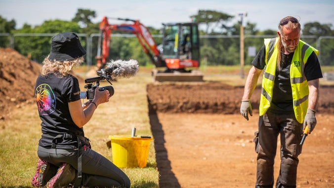 A female member of the Time Team crew wearing a branded t-shirt kneeling next to the trench, filming an archaeologist wearing a hi-vis at work in the trench